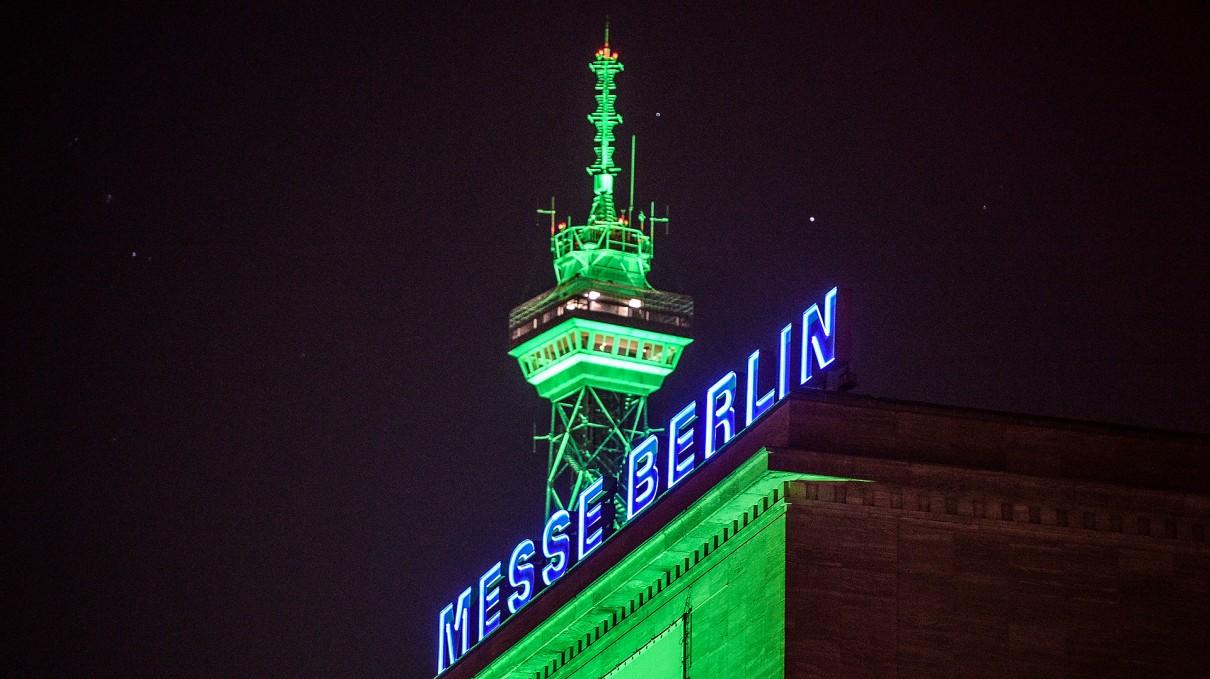 Im Hintergrund der grün beleuchtete Funkturm bei Nacht, im Vordergrund der beleuchtete Messe Berlin Schriftzug.